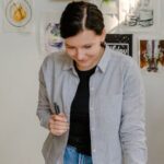 Choosing Artwork - Cheerful young female designer in jeans standing near wall with pictures and table with stationery while choosing drafts with letter design in daylight
