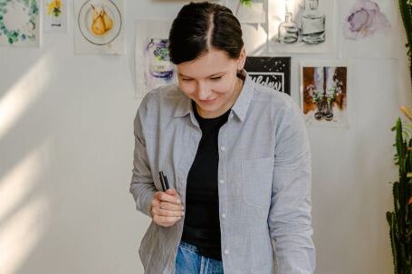 Choosing Artwork - Cheerful young female designer in jeans standing near wall with pictures and table with stationery while choosing drafts with letter design in daylight