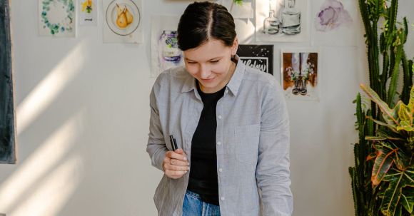 Choosing Artwork - Cheerful young female designer in jeans standing near wall with pictures and table with stationery while choosing drafts with letter design in daylight