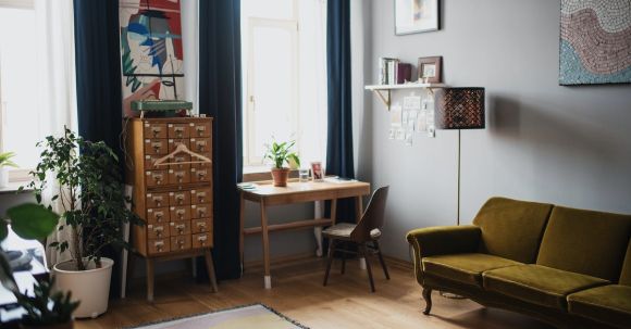 Interior Design - Comfortable couch put on wooden floor covered with rug and potted plants in apartment with big windows and dark curtains in sunlight