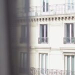 Neighborhood - Hotel with elegant balconies and bay windows in Paris as seen from opposite building through slightly ajar window curtains on sunny day