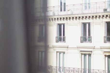 Neighborhood - Hotel with elegant balconies and bay windows in Paris as seen from opposite building through slightly ajar window curtains on sunny day