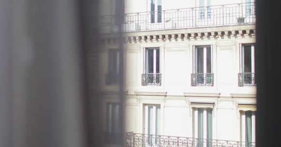 Neighborhood - Hotel with elegant balconies and bay windows in Paris as seen from opposite building through slightly ajar window curtains on sunny day