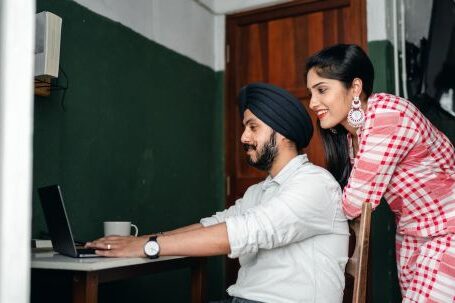 Home Office - Side view of positive young Sikh man in shirt and turban working on laptop at home while wife leaning on chair behind