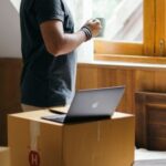 Neighborhood - Side view of young ethnic male owner of newly bought house with cup of coffee in bedroom full of boxes and suitcases standing and looking at neighboring houses through window