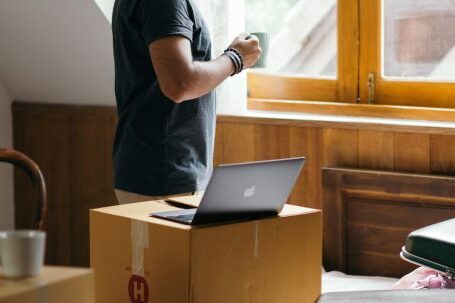 Neighborhood - Side view of young ethnic male owner of newly bought house with cup of coffee in bedroom full of boxes and suitcases standing and looking at neighboring houses through window