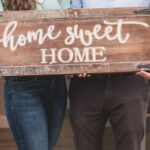 New Home - Happy Couple Standing Face to Face and Holding a Home Sweet Home Wooden Signage