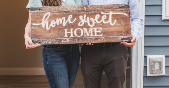 New Home - Happy Couple Standing Face to Face and Holding a Home Sweet Home Wooden Signage