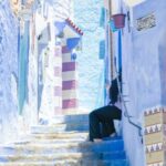 Neighborhood - Crop distant person sitting on colorful stairs between blue walls of buildings in sunny street in old town town of Morocco
