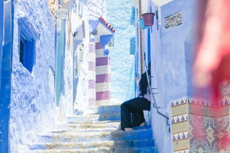 Neighborhood - Crop distant person sitting on colorful stairs between blue walls of buildings in sunny street in old town town of Morocco