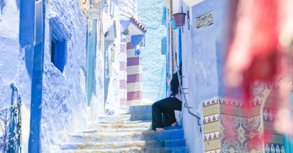Neighborhood - Crop distant person sitting on colorful stairs between blue walls of buildings in sunny street in old town town of Morocco